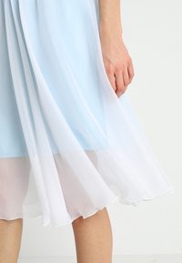 Vaaleansininen mekko, jonka leikkaus on kellotettu ja joka on valmistettu läpikuultavasta kankaasta. Helma muuttuu valkoiseksi, mikä korostaa sen kerroksellista ilmettä.