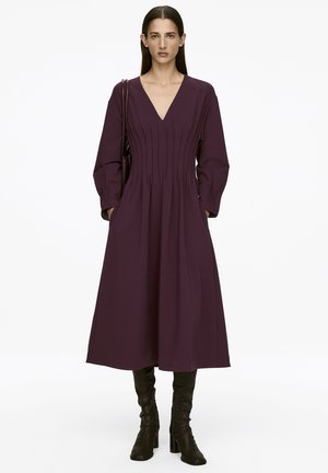 Langes lila Kleid mit langen Ärmeln, V-Ausschnitt, gefälteltem Detail, seitlichen Taschen und fließendem Saum, kombiniert mit schwarzen kniehohen Stiefeln.