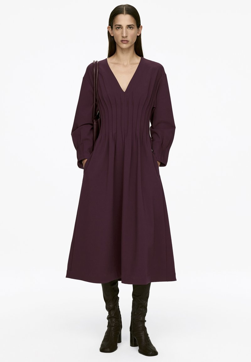 Langes lila Kleid mit langen Ärmeln, V-Ausschnitt, gefälteltem Detail, seitlichen Taschen und fließendem Saum, kombiniert mit schwarzen kniehohen Stiefeln.