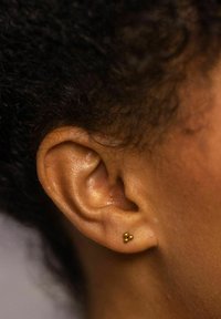 Goldene Ohrstecker mit drei kleinen Kugeln, vor einem Nahaufnahme von brauner Haut und lockigem Haar, das die Form und Textur des Ohrläppchens zeigt.