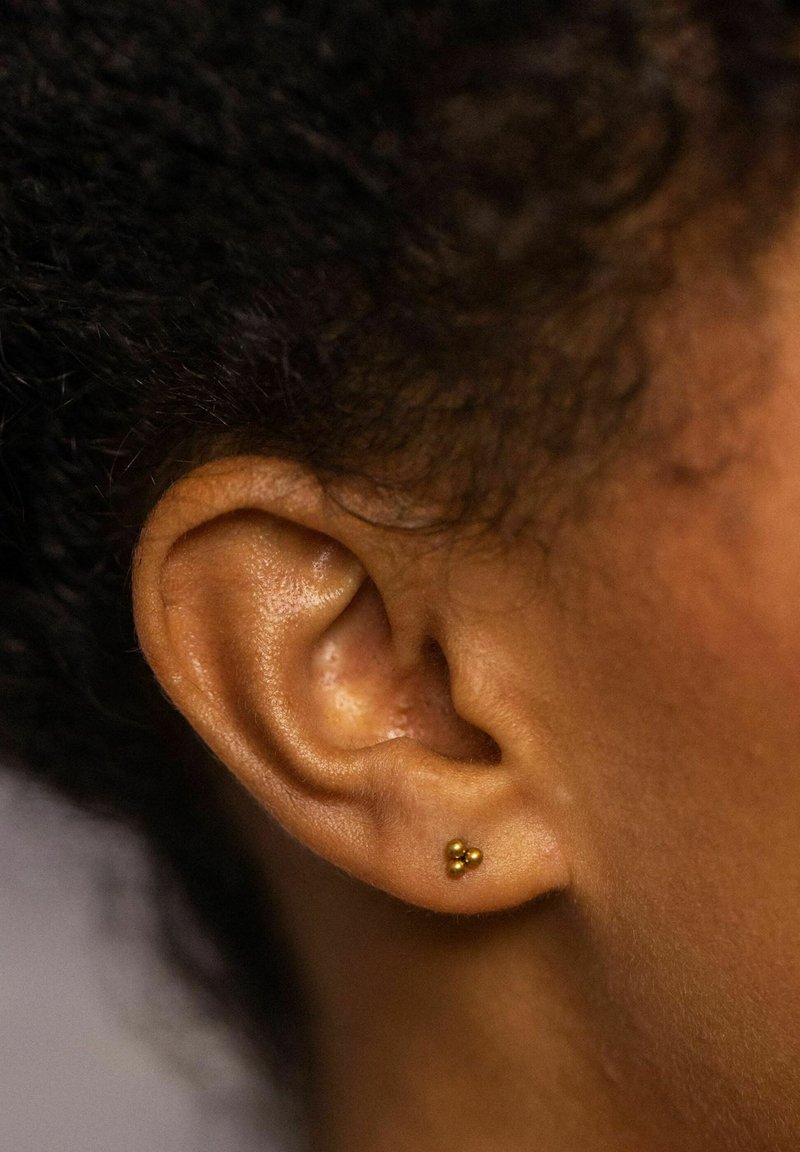 Goldene Ohrstecker mit drei kleinen Kugeln, vor einem Nahaufnahme von brauner Haut und lockigem Haar, das die Form und Textur des Ohrläppchens zeigt.
