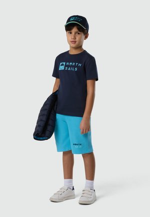 Garçon portant une casquette et un t-shirt North Sails bleu marine, un short North Sails turquoise, des baskets blanches, tenant une veste bleue marine dans une main.