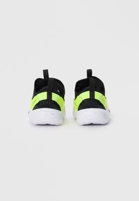 Une paire de baskets noires et jaune fluo avec des semelles blanches tachetées, vues de l'arrière sur un fond blanc uni.