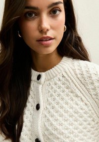 Kardigan w kolorze ecru, dziany, o strukturze wzoru, z okrągłym dekoltem i czarnymi guzikami. Posiada ściągacze przy mankietach oraz miękką fakturę.