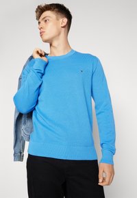 Niebieski sweter z dzianiną waffle, z okrągłym dekoltem i ściągaczami przy mankietach. Naszywka z logo na piersi. Model trzyma dżinsową kurtkę na jednym ramieniu.