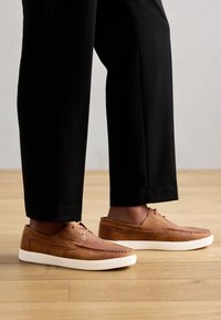 Hnedé kožené topánky s bielymi podrážkami, s jednoduchým dizajnom šnurovania, detailmi šitia a hladkým povrchom, skombinované s čiernymi nohavicami.