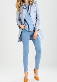 Manteau trench bleu clair avec des boutons noirs, superposé à un haut en denim et un jean skinny bleu. Fini avec des chaussures à talons hauts orange.