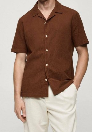 Mann trägt braunes Kurzarmhemd mit Knöpfen und hellbeige Hose, eine Hand in der Tasche, vor einfarbigem Hintergrund.