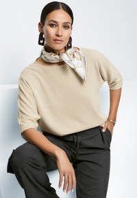 Beige gebreide trui met structuur en korte mouwen, gecombineerd met donkere, op maat gemaakte broek. Een dessin sjaal accentueert de halslijn.