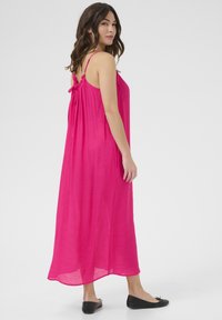 Robe maxi fuchsia avec des fines bretelles, détail noué froncé au dos, tissu fluide et coupe relaxante, associée à des ballerines noires.