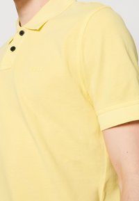 Personne portant un polo BOSS jaune clair avec deux boutons noirs, manches courtes et col, cadré au niveau du cou et du bras.
