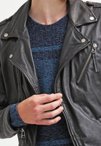 Czarna skórzana kurtka motocyklowa ze srebrnymi elementami, zamkami błyskawicznymi i zatrzaskami. Nałożona na niebieski, teksturowany sweter z dzianiny.