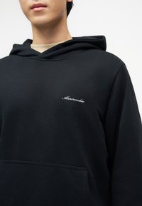 Νέος/νέα που φοράει μαύρο φούτερ με κουκούλα και μπροστινή τσέπη, με λευκό καλλιγραφικό λογότυπο "Abercrombie" στο στήθος, μπροστά σε απλό φόντο.