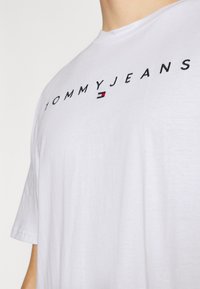 Balta medvilninė marškinėlių su "TOMMYJEANS" logotipu juodu siuvinėjimu, mažas raudonas, baltas ir mėlynas vėliavos akcentas. Trumpomis rankovėmis.