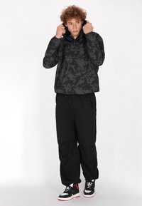 Jachetă neagră cu glugă, cu un model geometric texturat, asortată cu pantaloni negri largi și teniși negri cu accente roșii.