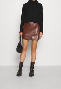 Svart stickad polotröja, brun imitationläder mini kjol med textur, och knähöga bruna stövlar. Svart handväska i handen, stående mot en grå bakgrund.