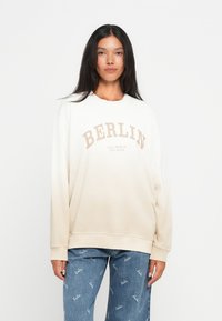 Sweatshirt em degradê bege com a impressão "BERLIN" em castanho, gola redonda e punhos canelados. Combinado com jeans azuis com um padrão de logotipo repetido.