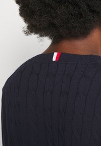 Maglione lavorato a maglia blu navy con un motivo a trecce e un accento rosso e bianco nella parte posteriore del collo. La texture è a costine e leggermente spessa.