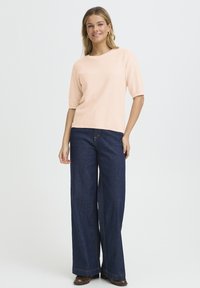 Gaiši rozā īss piedurkņu tops ar teksturētu apdari, kopā ar tumši zilām džinsa platām biksēm, kurām ir redzama šuve pa vidu priekšpusē.