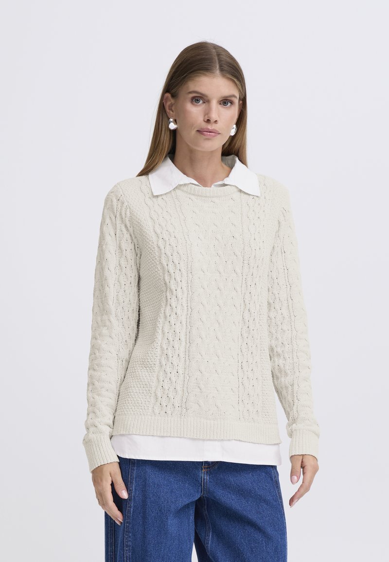 Maglione beige chiaro a trecce con collo rotondo a coste, caratterizzato da motivi testurizzati, indossato sopra una camicia bianca con colletto, abbinato a jeans blu.