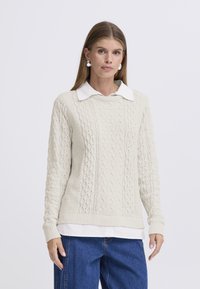 Light beige cable-knit sweater with a ribbed crew neck, featuring textured patterns, worn over a white collared shirt, paired with blue jeans.