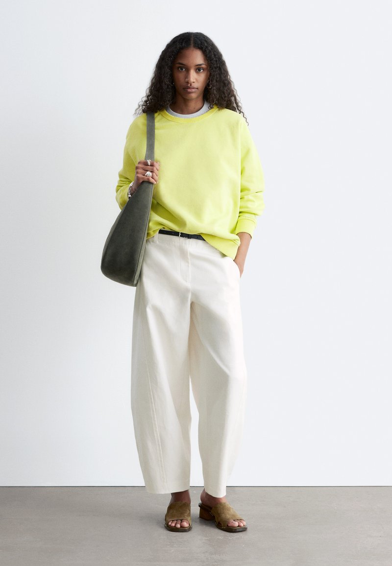 Femme aux cheveux bouclés portant un sweat jaune, un large pantalon blanc, des sandales marron et un sac à bandoulière vert foncé.