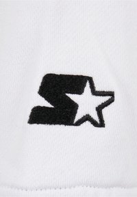 Witte stof heeft een zwart geborduurd logo met een gestileerde "S" en een ster. De textuur is glad met een gebreid patroon.