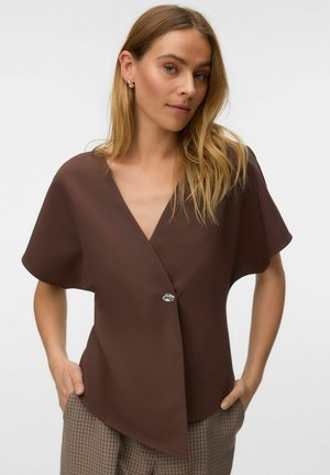 Femme aux cheveux blonds portant un chemisier marron ample à col en V avec des manches courtes et un pantalon à carreaux, mains dans les poches, fond neutre.