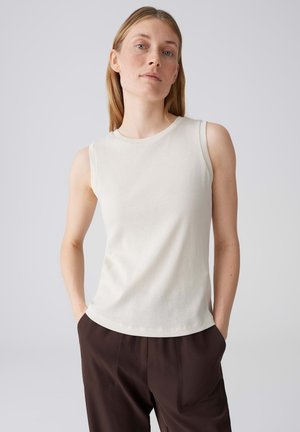 Femme aux cheveux raides portant un haut blanc sans manches et un pantalon marron foncé, les mains dans les poches, devant un fond uni.