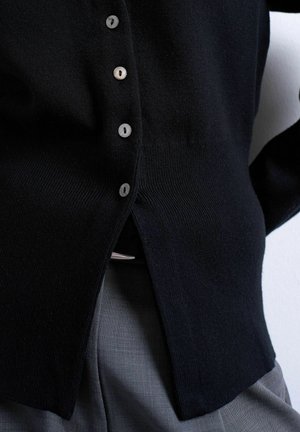 Zwart gebreid vest met knopen, ingestopt in grijze broek met zichtbare riem gesp, met focus op de taille en kledingdetails.