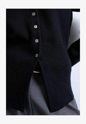 Black buttoned cardigan tucked into gray pants with a visible belt buckle, focusing on midsection and clothing details.
