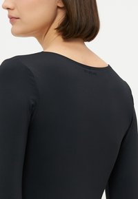 Črn bodysuit z dolgimi rokavi, ki vsebuje gladek in raztegljiv material ter širok, zaobljen izrez. Ime blagovne znamke je subtilno vtisnjeno na hrbtu.