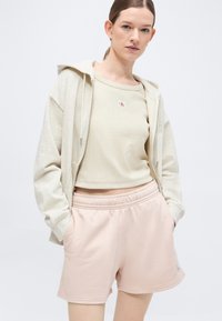 Νεαρή γυναίκα που φοράει μπεζ φωλιαστό φούτερ με φερμουάρ, κρεμ κοντό τοπ με μικρό λογότυπο και ανοιχτό ροζ σορτς με ελαστική μέση, με τα χέρια στις τσέπες.