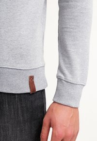 Jasnoszara bluza z prążkowanymi mankietami. Zawiera brązową skórzaną metkę przy dolnym brzegu. Tkanina wydaje się miękka o gładkiej fakturze.