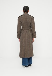 Long plaid trench coat with a belt, featuring brown, beige, and red tones; designed with a pointed collar and wide sleeves. Denim flared pants below.