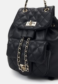 Sac à dos en cuir noir avec motif matelassé, bretelles en chaîne et une boucle dorée. Comprend une poche avant avec un rabat distinctif.