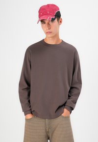 Cămașă maro cu mânecă lungă, guler rotund și croială lejeră. Împerecheată cu pantaloni bej deschis cu un model și o șapcă roșie cu negru.