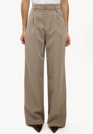 Pantalons beiges taille haute avec des rayures blanches verticales, des plis, des jambes larges et une texture lisse, associés à des chaussures pointues marron.