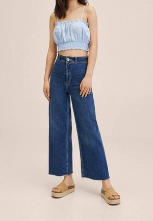 Kobieta w jasno-niebieskim, marszczonym topie z odsłoniętym brzuchem, wysokim stanem szerokich niebieskich dżinsach oraz beżowych sandałach na platformie, stojąca na neutralnym tle.