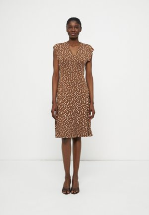 Γυναίκα που στέκεται, φοράει καφέ φόρεμα μέχρι τα γόνατα με λευκές πουά και μαύρα ανοιχτά παπούτσια με τακούνι, σε απλό λευκό φόντο.