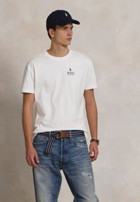 Jauns vīrietis, ģērbies balts Polo Ralph Lauren T-kreklā, nolietotās zilās džinsēs, svītrainā auduma jostā, melnā cepurē un ar rokas pulksteni, skatās uz sāniem.