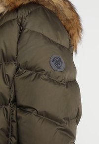 Giacca a vento verde con design trapuntato, dotata di cappuccio con bordo in pelliccia, una patch circolare nera e una texture lucida e liscia.