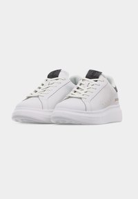 Ζεύγος λευκών αθλητικών παπουτσιών χαμηλού ύψους με μαύρες πίσω λωρίδες, κορδόνια και χοντρές λευκές σόλες, παρουσιασμένα σε απλό φωτεινό φόντο.