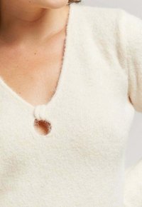 Pull en tricot crème avec un décolleté en V profond et une petite découpe circulaire accentuée par un nœud. Tissu doux et texturé.