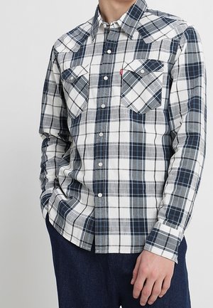 Homme portant une chemise à manches longues à carreaux blanc, gris et noir boutonnée, avec deux poches poitrine, associée à un pantalon foncé.