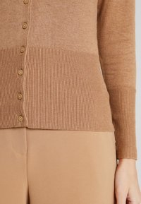 Braune Strick-Cardigan mit langen Ärmeln und geripptem Saum, ausgestattet mit sechs hellbraunen Knöpfen auf der Vorderseite und einer glatten Textur.