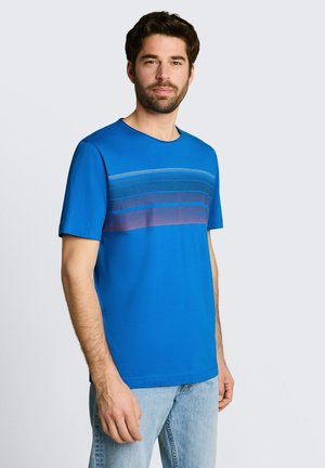 Mężczyzna z ciemnymi włosami i brodą, ubrany w niebieski krótki t-shirt w poziome paski na klatce piersiowej oraz jasnoniebieskie dżinsy.