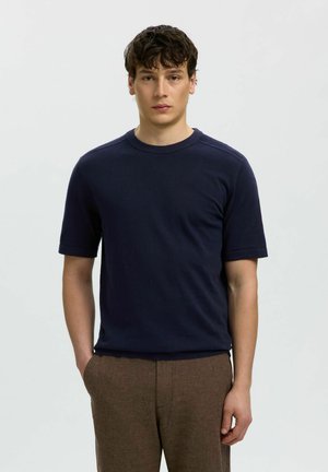 Joven con cabello rizado oscuro que lleva una camisa azul marino de manga corta y pantalones marrones, de pie con una mano en el bolsillo sobre un fondo liso.