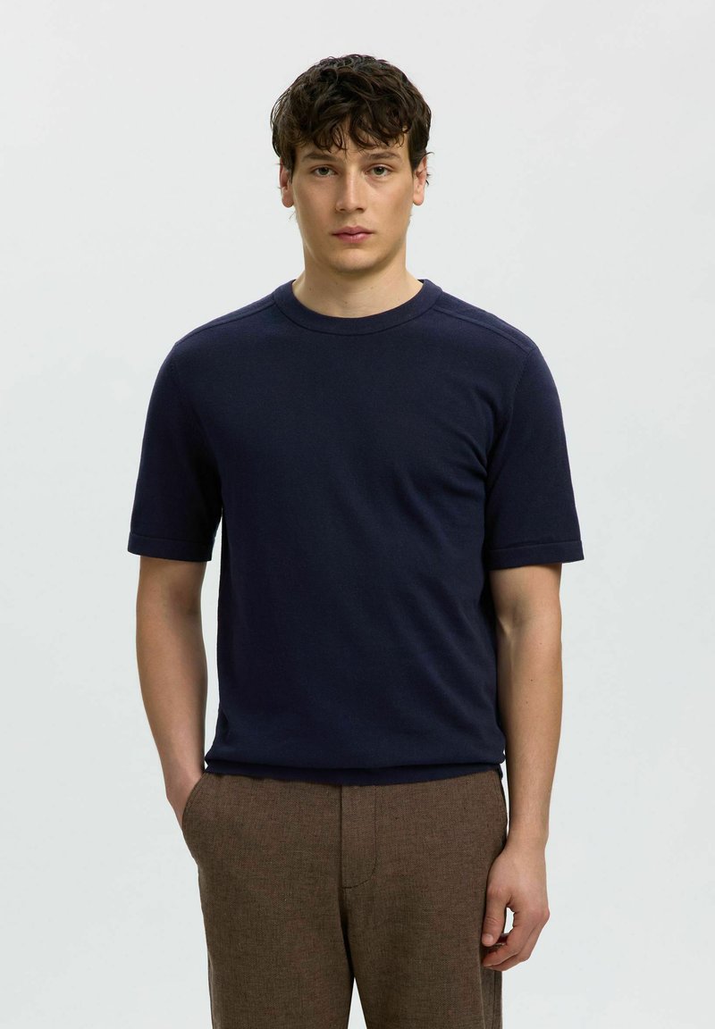 Jeune homme aux cheveux bouclés foncés portant une chemise bleu marine à manches courtes et un pantalon marron, debout avec une main dans la poche sur un fond uni.