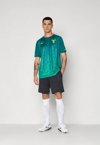 Žalios spalvos sportinis marškinėlis su geltonomis detalėmis, derinamas su juodais šortais. Modelis dėvi baltas kojines ir sportinius batelius, ant rankų matomos tatuiruotės.